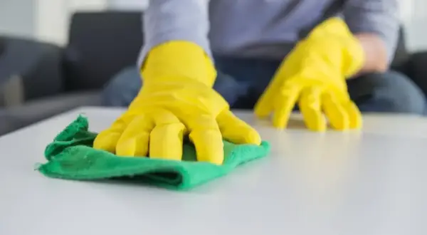 Manos con guantes amarillos limpiando una superficie blanca con un trapo verde. Aprende como lavar los trapos de limpieza para un hogar impecable.