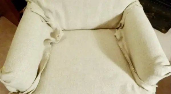 Tela beige drapeada y ajustada con alfileres en los brazos de una silla, parte de un tutorial sobre como hacer fundas para sillas.