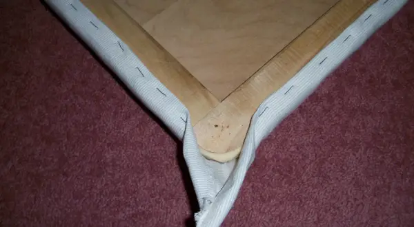Detalle de cómo tapizar esquinas de un bastidor de madera con tela blanca, asegurada con grapas.