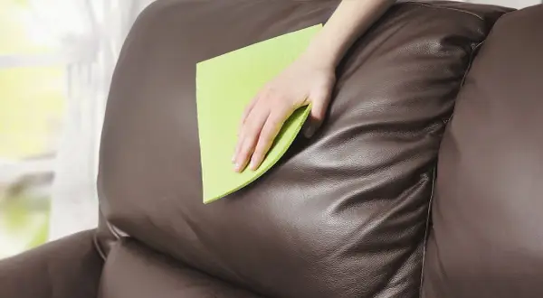 Mano limpiando un sofá de piel marrón con un paño verde. Aprende cómo limpiar sofá de piel para mantenerlo como nuevo.