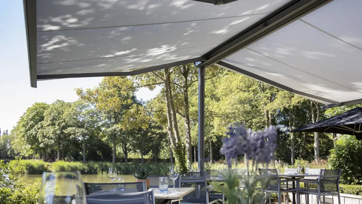 Terraza con mesas y sillas bajo una lona que es un toldo retráctil, con vistas a un río y árboles frondosos.