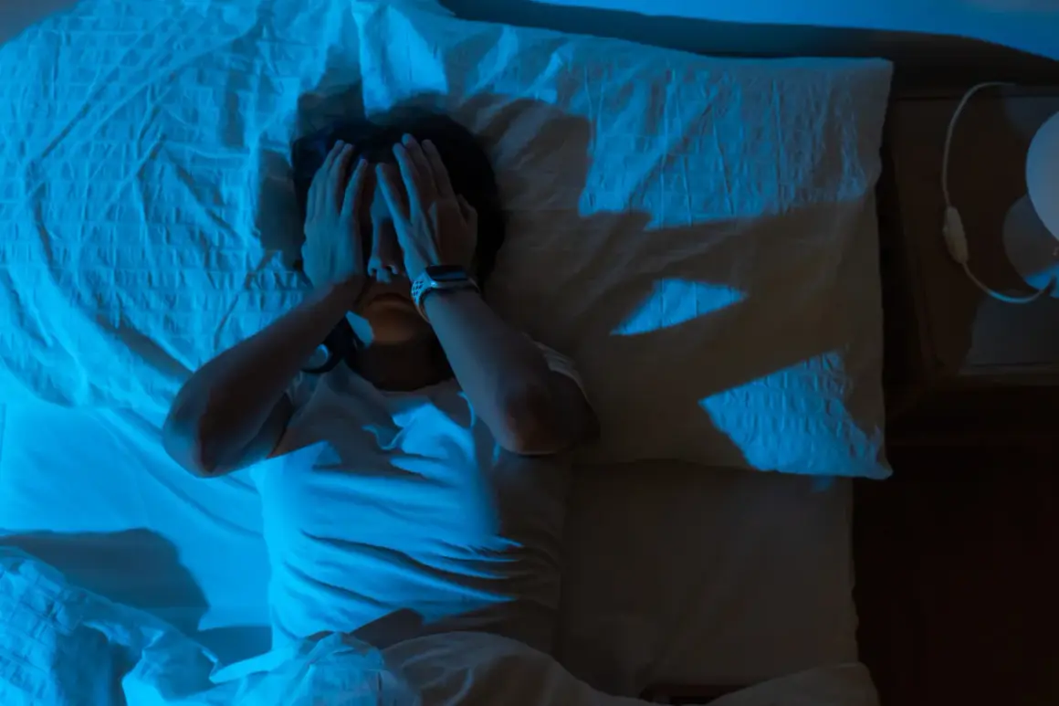 Persona en la cama, con las manos cubriendo su rostro, en un ambiente oscuro y con luz azul. Parece estar despertándose de golpe asustado.