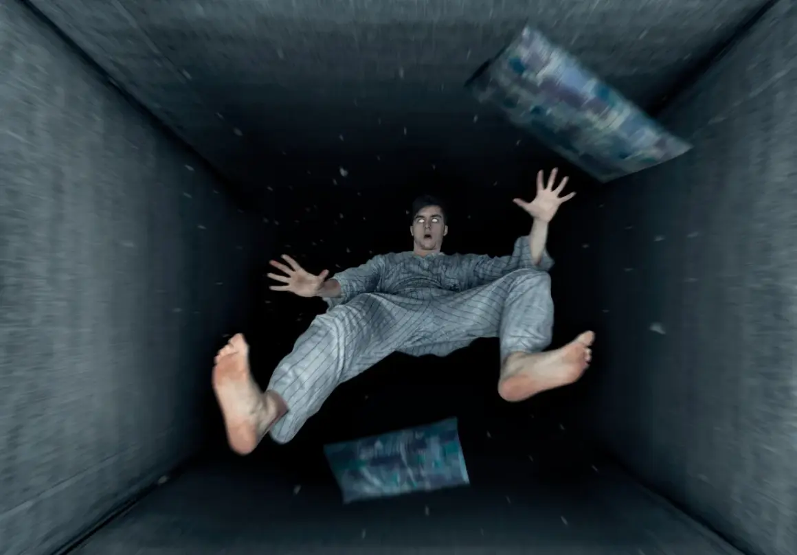 Un hombre en pijama experimenta una caida al vacio dentro de un túnel oscuro, con almohadas flotando a su alrededor.