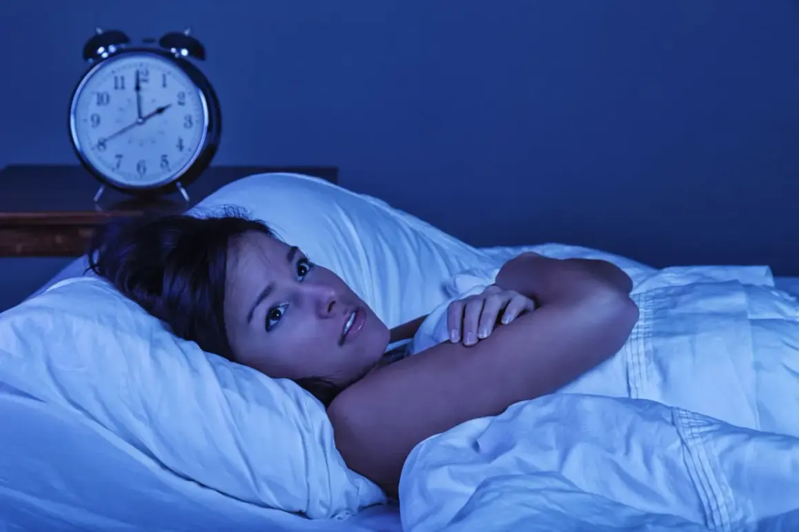 Mujer en la cama, con los ojos muy abiertos, sintiendo que se está despertando de golpe con taquicardia. Un despertador antiguo marca las 3:10 AM.