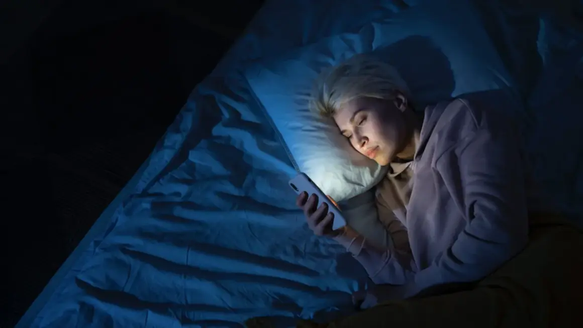 Mujer rubia en la cama, mirando su móvil con luz azul. Quizás por eso me despierto a las 4 de la mañana.