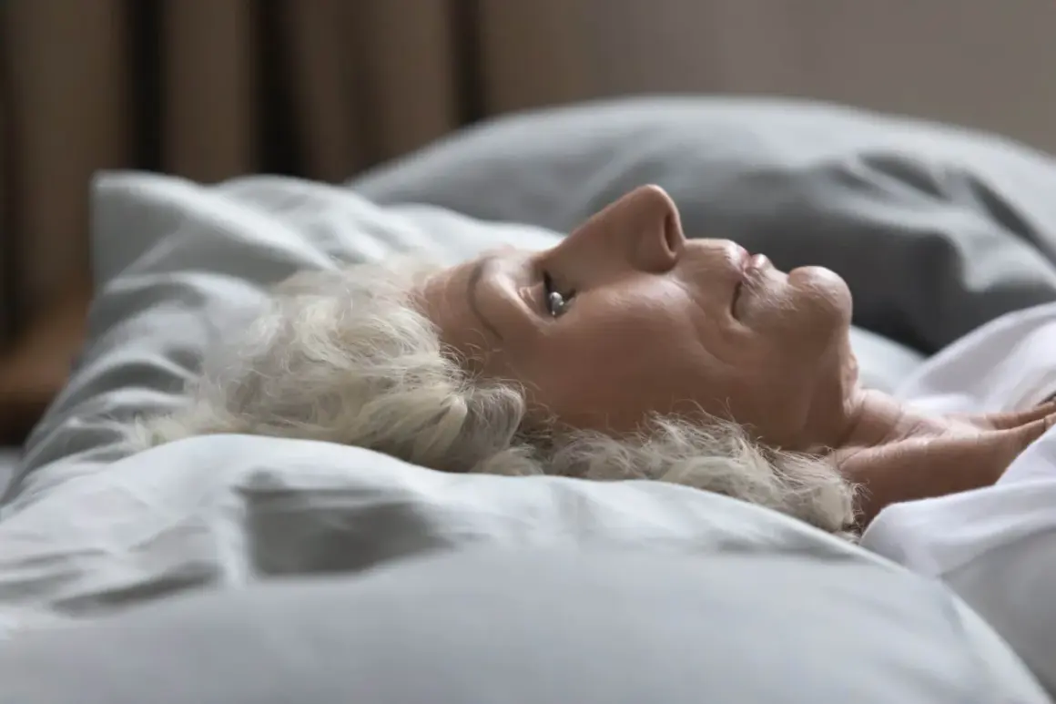 Mujer mayor con cabello blanco yace en la cama, con los ojos entreabiertos, como si soñara que duermo con los ojos abiertos.