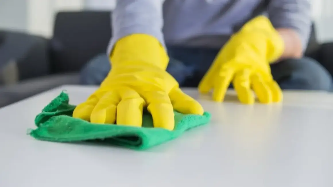 Manos con guantes amarillos limpiando una superficie blanca con un trapo verde. Aprende como lavar los trapos de limpieza para un hogar impecable.