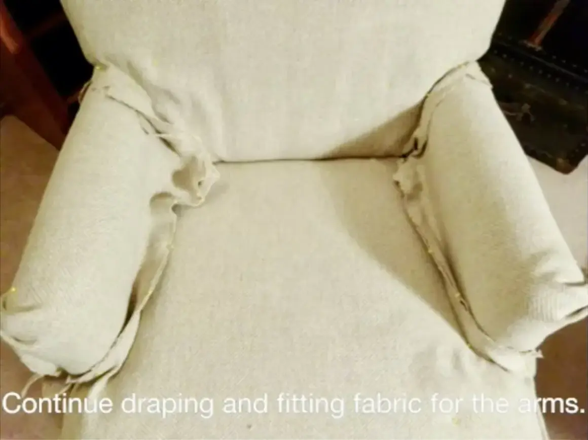 Tela beige drapeada y ajustada con alfileres en los brazos de una silla, parte de un tutorial sobre como hacer fundas para sillas.
