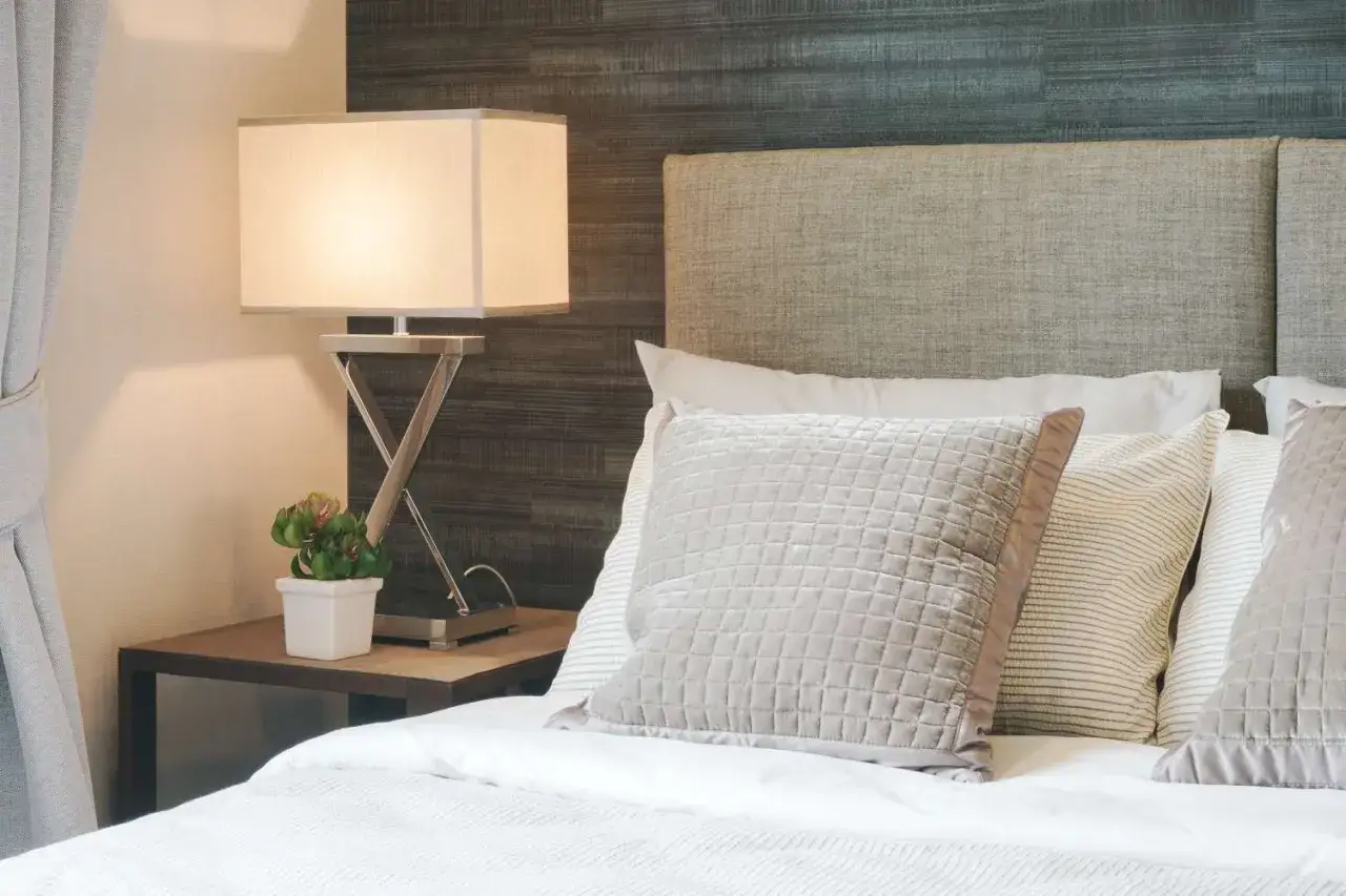 Cabecero de tela gris en cama con cojines, l&aacute;mpara y planta. Aprende como limpiar cabecero de tela para mantenerlo impecable.