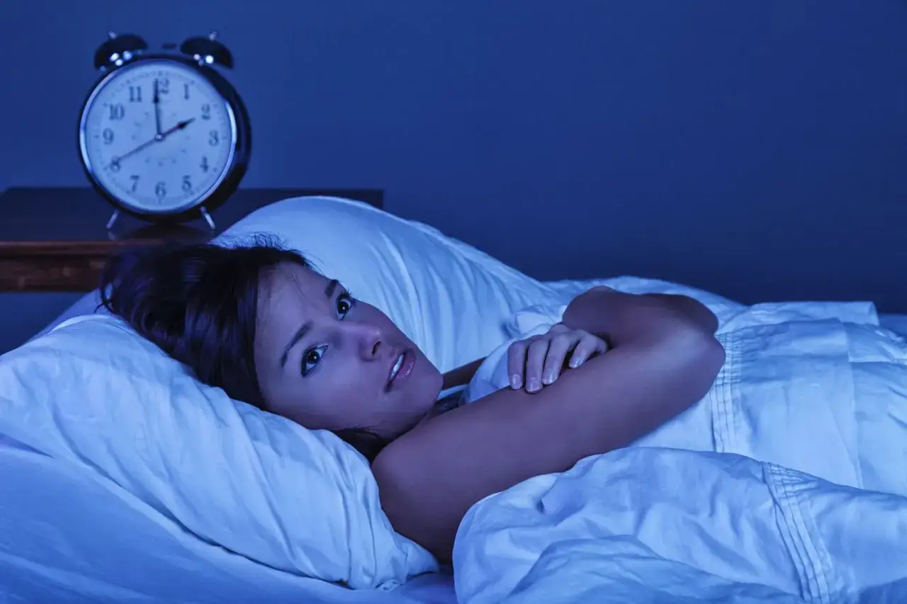Mujer en la cama, con ojos abiertos y expresi&oacute;n de angustia, despertarse de golpe con taquicardia. El despertador marca las 3:15 AM.