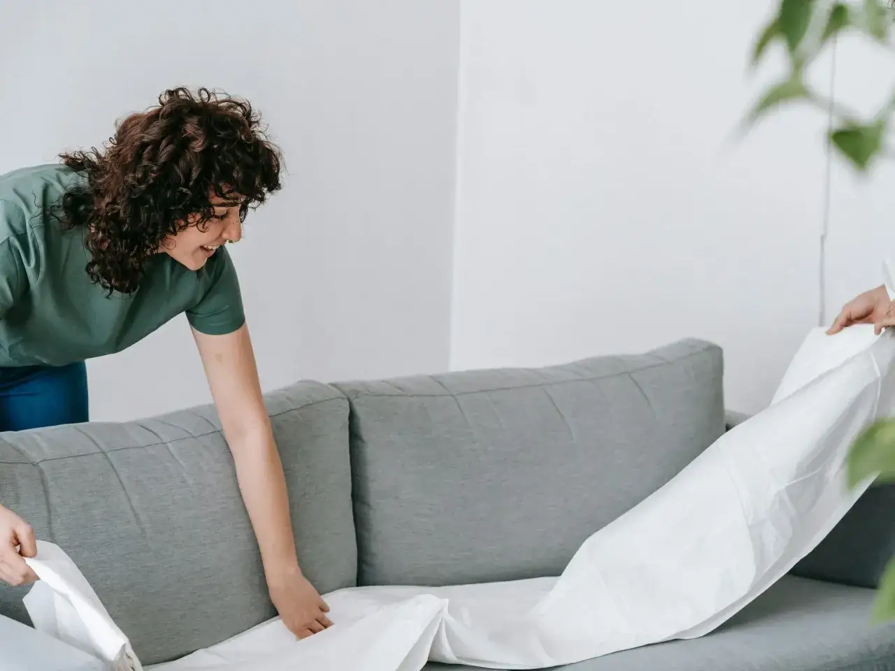 Dos personas colocan una funda blanca sobre un sof&aacute; gris. Una forma f&aacute;cil de saber como limpiar un sofa de tela sin aspiradora.