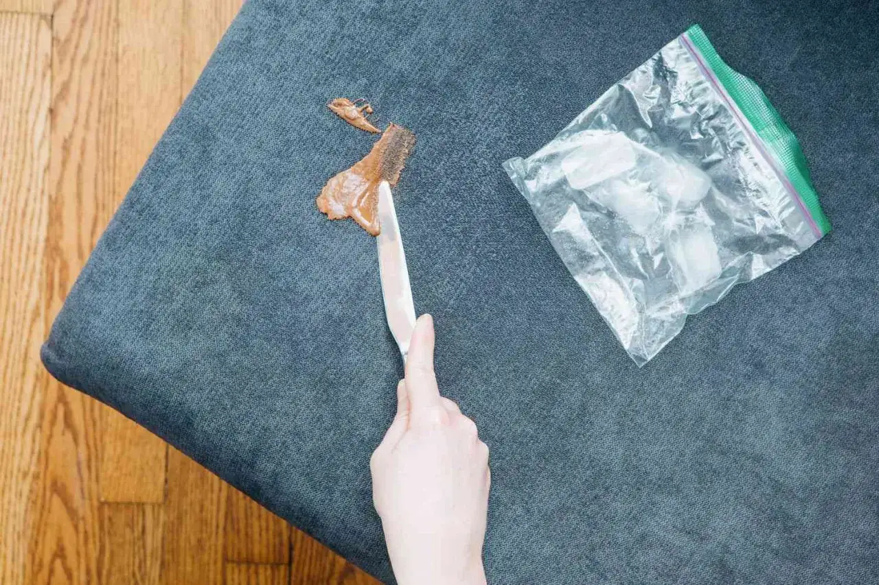 Mano raspando mancha de chocolate del sof&aacute; con cuchillo. Bolsa con hielo al lado, para saber como quitar manchas de chocolate del sofa.