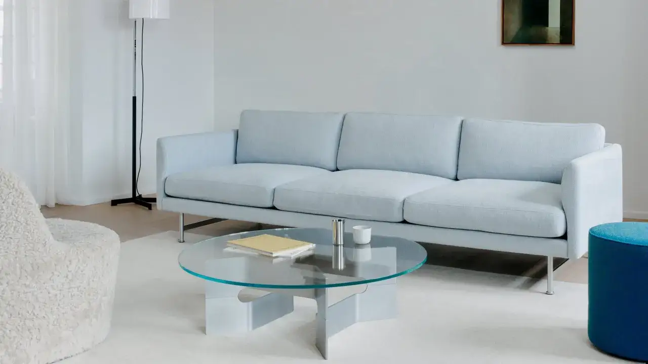 Un sof&aacute; azul claro con tres cojines. En el centro, una mesa de centro de cristal con un libro y una taza. A la izquierda, un sill&oacute;n blanco peludo.