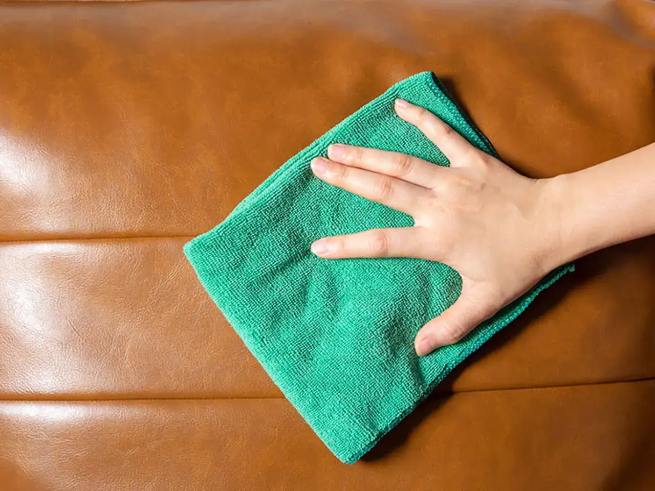 Mano limpiando un sofá de piel marrón con un paño de microfibra verde. Guía para como limpiar sofa de piel.