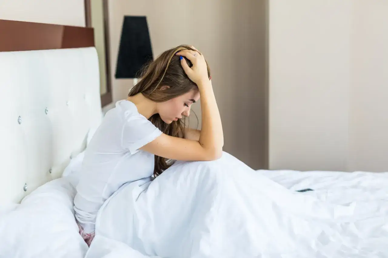 Una joven se sienta en la cama, abrazándose las rodillas, con la cabeza entre las manos. Parece que ha estado despertarse de golpe asustado.