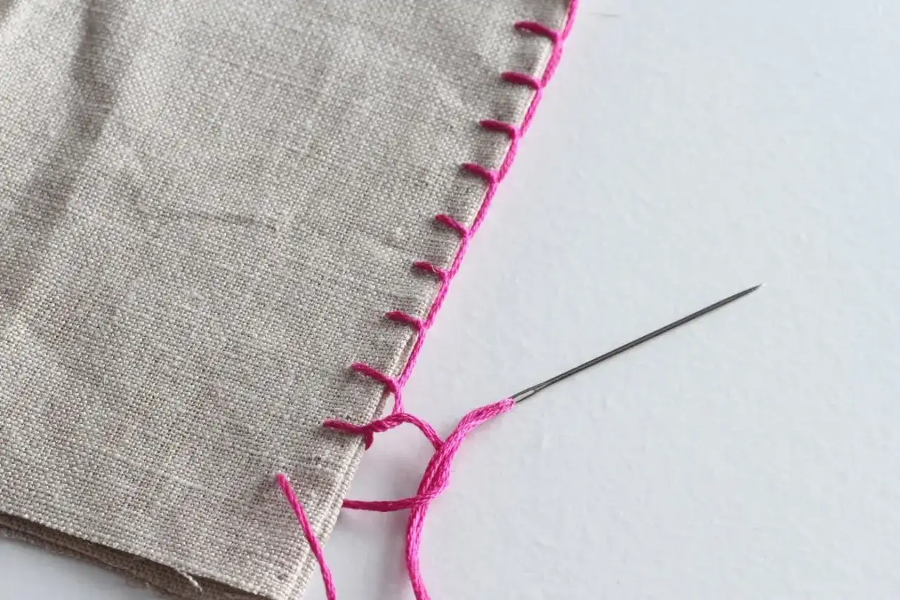 Detalle de puntadas rosas para rematar bordes de tela a mano. Una aguja con hilo rosa está lista para continuar el trabajo.