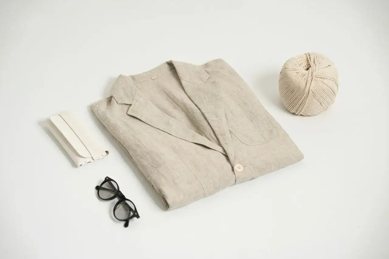 Un blazer de lino beige doblado, junto a gafas, un estuche y una bola de hilo. Ideal para aprender como encoger el lino.