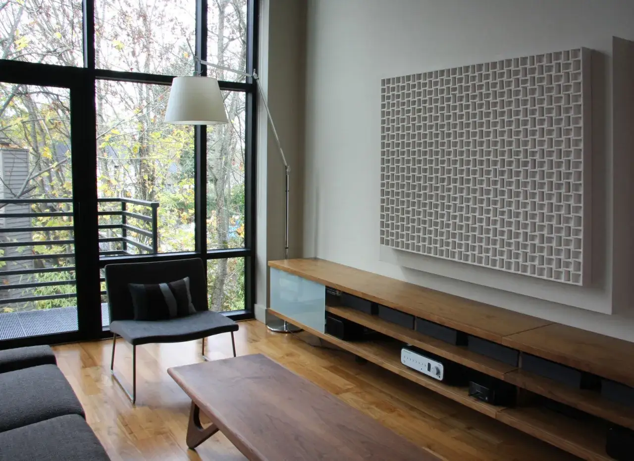 Un panel decorativo en la pared, que parece hecho de fieltro, complementa un mueble de madera y un sill&oacute;n junto a un gran ventanal.