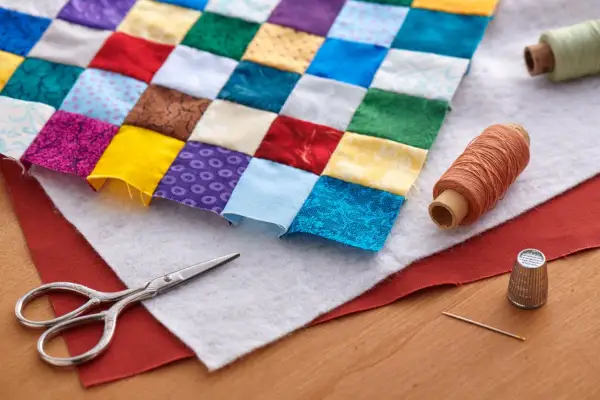 Un trozo de tela de patchwork con cuadrados de colores, tijeras, hilos y dedal. Así se empieza un proyecto de patchwork.