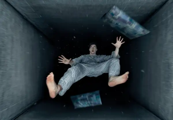 Un hombre en pijama experimenta una caida al vacio dentro de un túnel oscuro, con almohadas flotando a su alrededor.