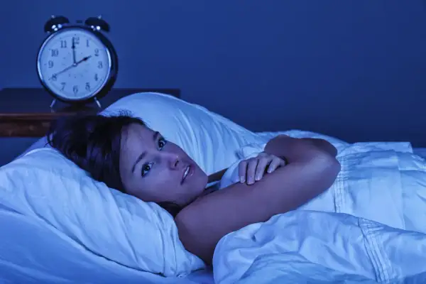 Mujer en la cama, con los ojos muy abiertos, sintiendo que se está despertando de golpe con taquicardia. Un despertador antiguo marca las 3:10 AM.