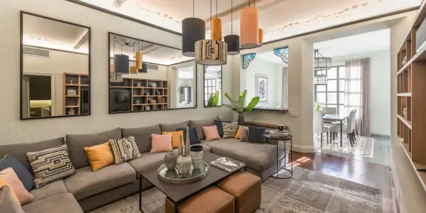 Amplios salones con sofás modulares grises, cojines coloridos y una mesa de centro. Reflejos en espejos y lámparas colgantes añaden estilo.