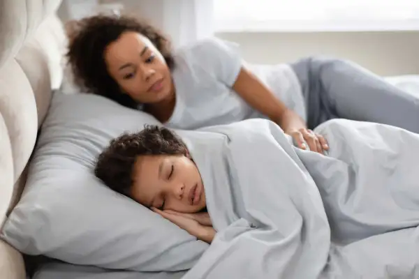Madre observa a su hijo dormir plácidamente. Quizás se pregunta por qué se habla en sueños, o simplemente disfruta de la paz del momento.