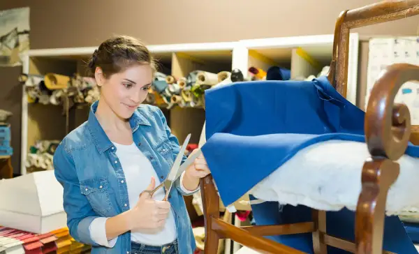 Mujer cortando tela azul para como tapizar un sillón pequeño. Estanterías con rollos de tela al fondo.