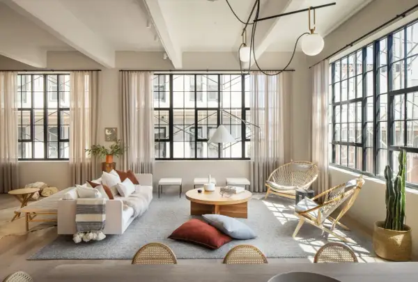 Un salón estilo industrial nórdico con sofá blanco, mesas de centro de madera, sillas de ratán y grandes ventanales.